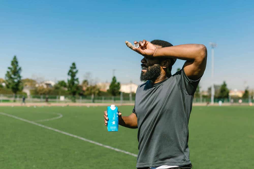 heat-related illness - man on sunny soccer field, shielding eyes from sun, drinking water